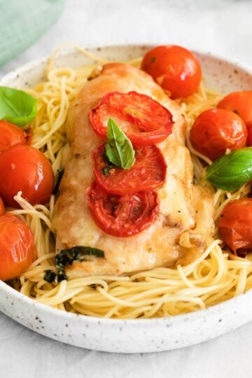 Square format close-up of a serving of Chicken Margherita on a bed of spaghetti, surrounded by red cherry tomatoes and green basil leaves. The chicken breast is topped with sliced tomatoes and cheese.