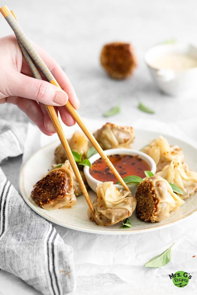 A hand holding chopsticks is picking up one of the mushroom wonton dumplings from a white plate surrounded by dipping sauce and fresh basil leaves.