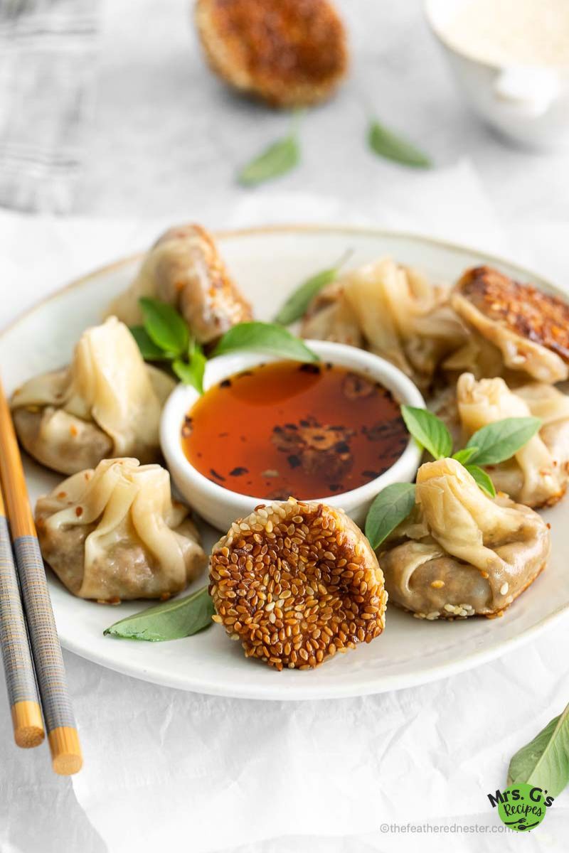 A plate of pan-fried mushroom dumplings arranged around a central bowl of chili oil dipping sauce, garnished with fresh basil leaves. One dumpling is flipped to show its golden, sesame-crusted bottom.