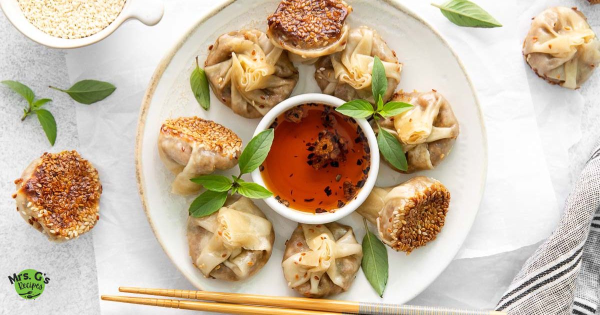 Overhead shot of a white plate holding seven mushroom wonton dumplings (some pan-fried with sesame seeds on the bottom, others folded like money bags) arranged around a small bowl of chili oil dipping sauce.