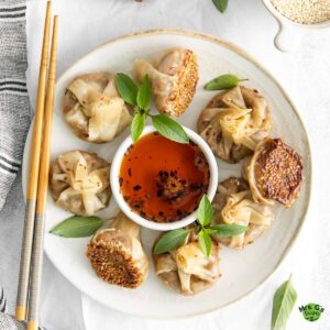 Square-cropped overhead photo of eight mushroom wonton dumplings arranged on a plate around a small white bowl of chili oil, with bamboo chopsticks resting alongside.