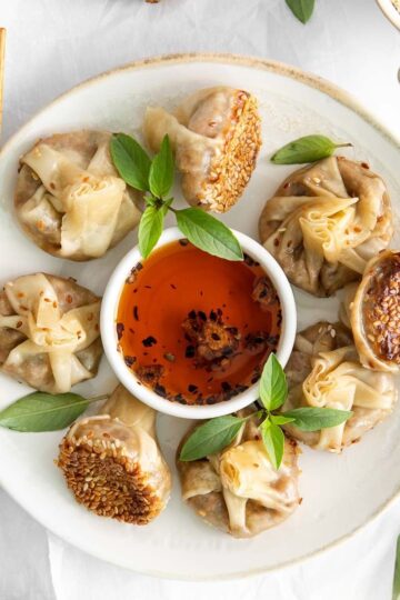 Square-cropped overhead photo of eight mushroom wonton dumplings arranged on a plate around a small white bowl of chili oil, with bamboo chopsticks resting alongside.