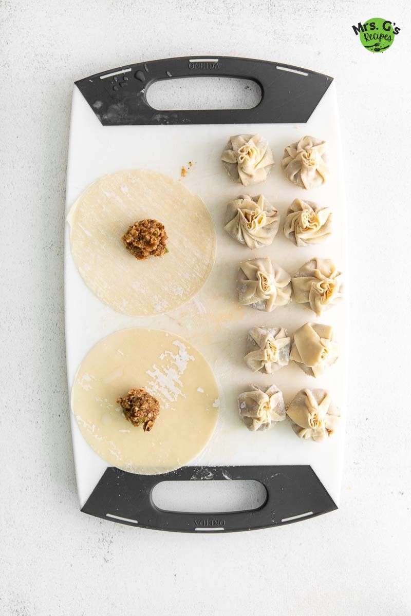 Overhead view of a white cutting board showing the dumpling assembly process: two round wonton wrappers are topped with a spoonful of savory filling, and several completed, uncooked wonton money bag dumplings are lined up next to them.