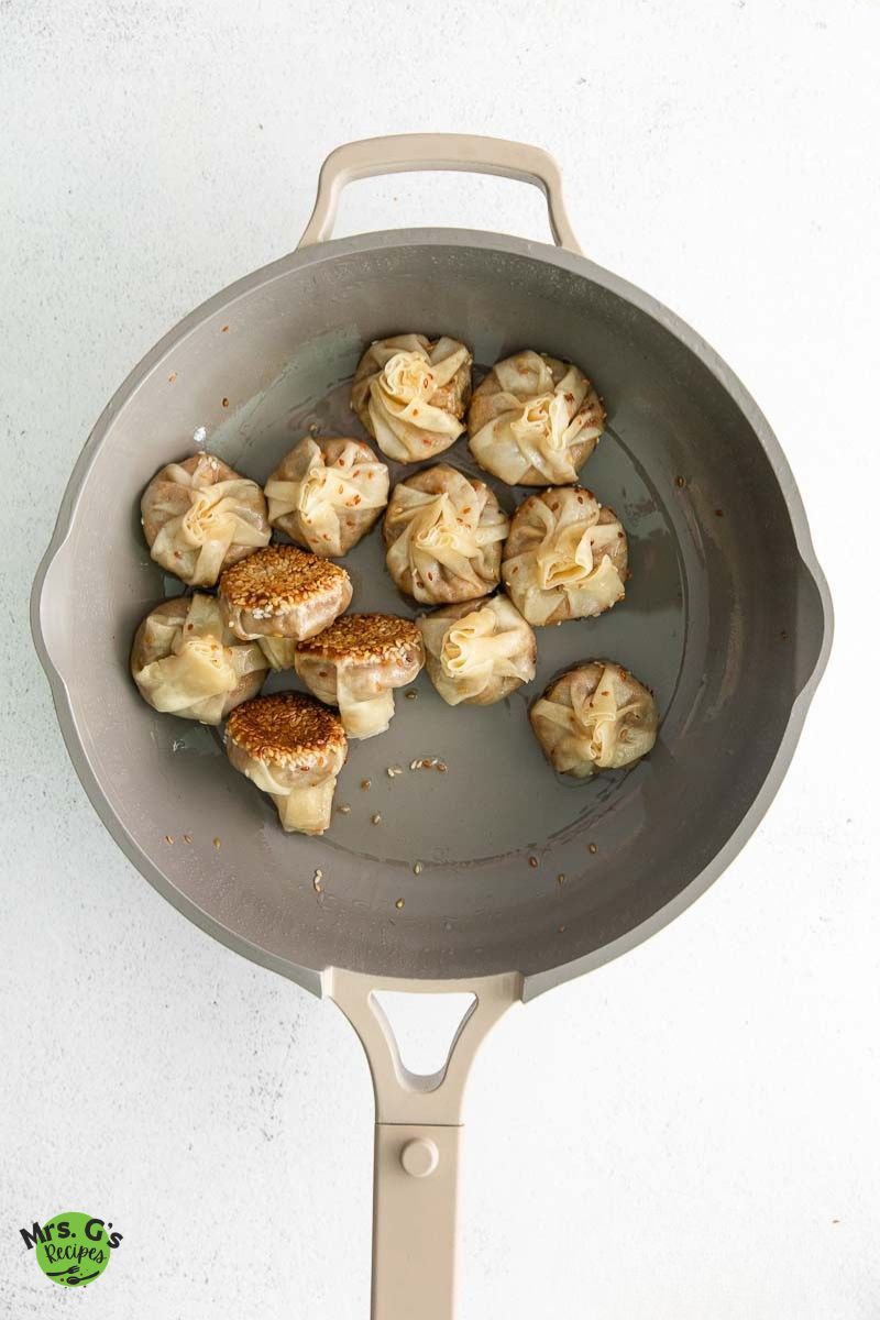 Overhead view of about a dozen partly cooked wonton dumplings in a gray non-stick skillet. Some are lightly seared and coated with sesame seeds on the bottom.