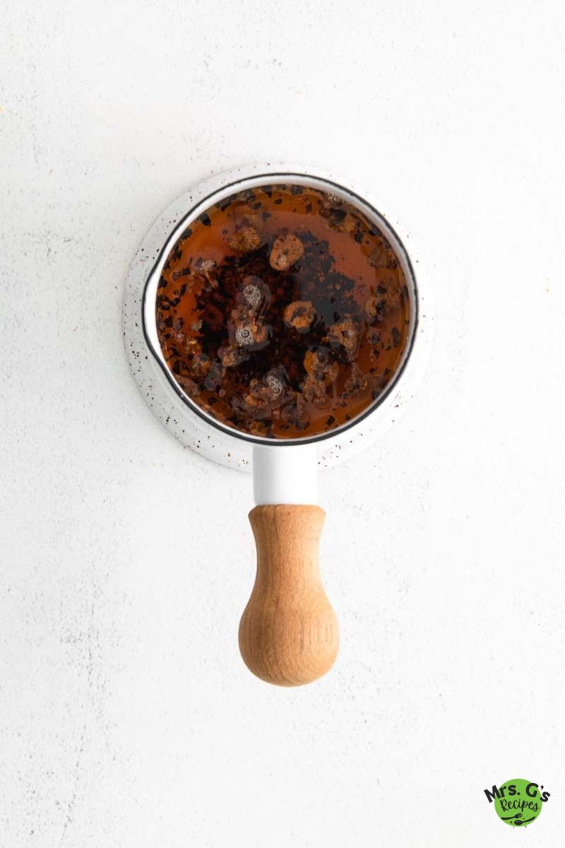 Top-down view of a small white saucepan with a wooden handle, containing a dark, oily chili dipping sauce with visible spices and bits of seasoning.