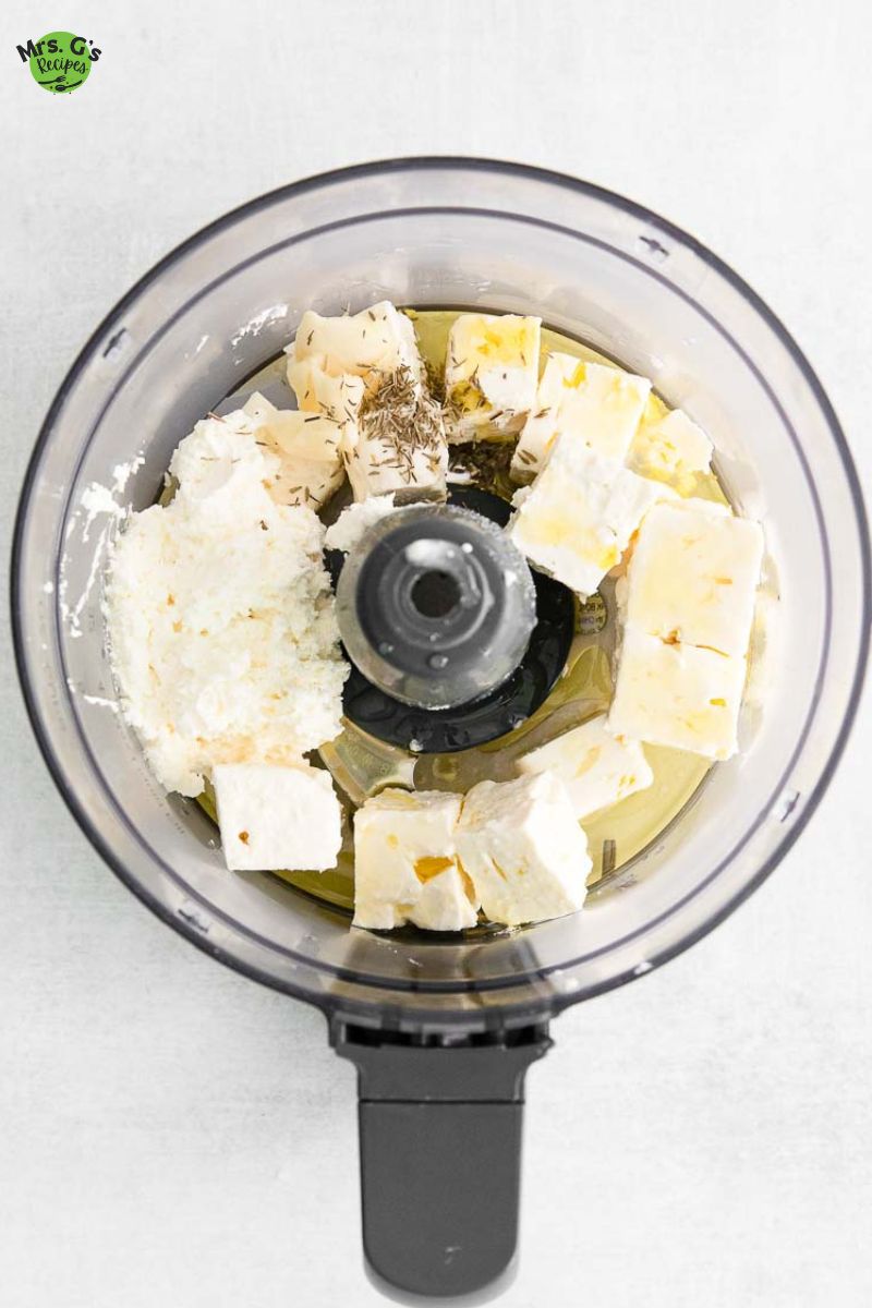 a food processor with feta, cream cheese, olive oil, garlic, lemon juice, and thyme in it.