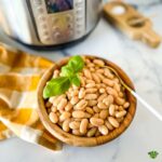 A wooden bowl of pressure cooked great northern beans with a sprig of basil on top and a gold and white napkin and instant pot in the background.