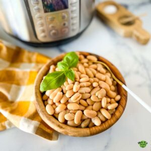 A wooden bowl of pressure cooked great northern beans with a sprig of basil on top and a gold and white napkin and instant pot in the background.