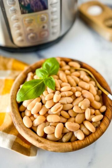 A wooden bowl of pressure cooked great northern beans with a sprig of basil on top and a gold and white napkin and instant pot in the background.