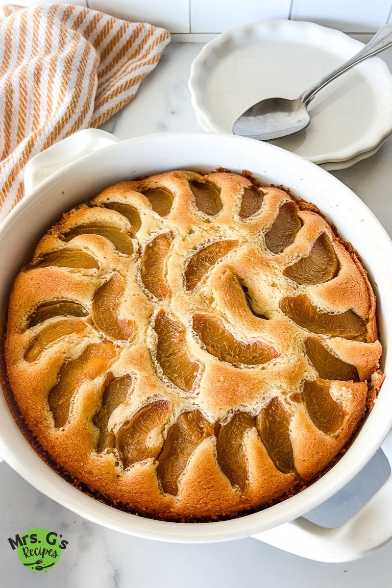 Peach cobbler pound cake baked in a round white baking dish. Stack of dessert plates and spoons sitting behind the pound cake.