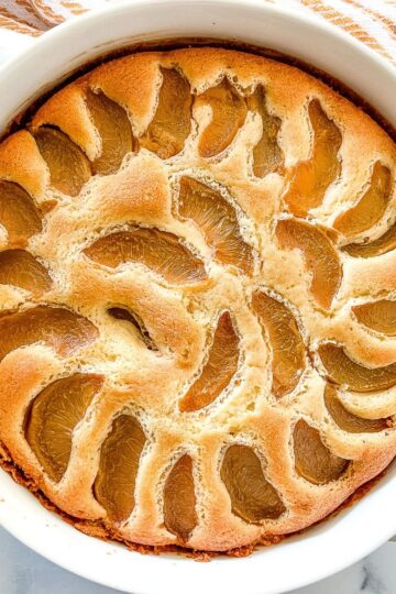 Peach cobbler pound cake baked in a round white baking dish. Stack of dessert plates and spoons sitting behind the pound cake.