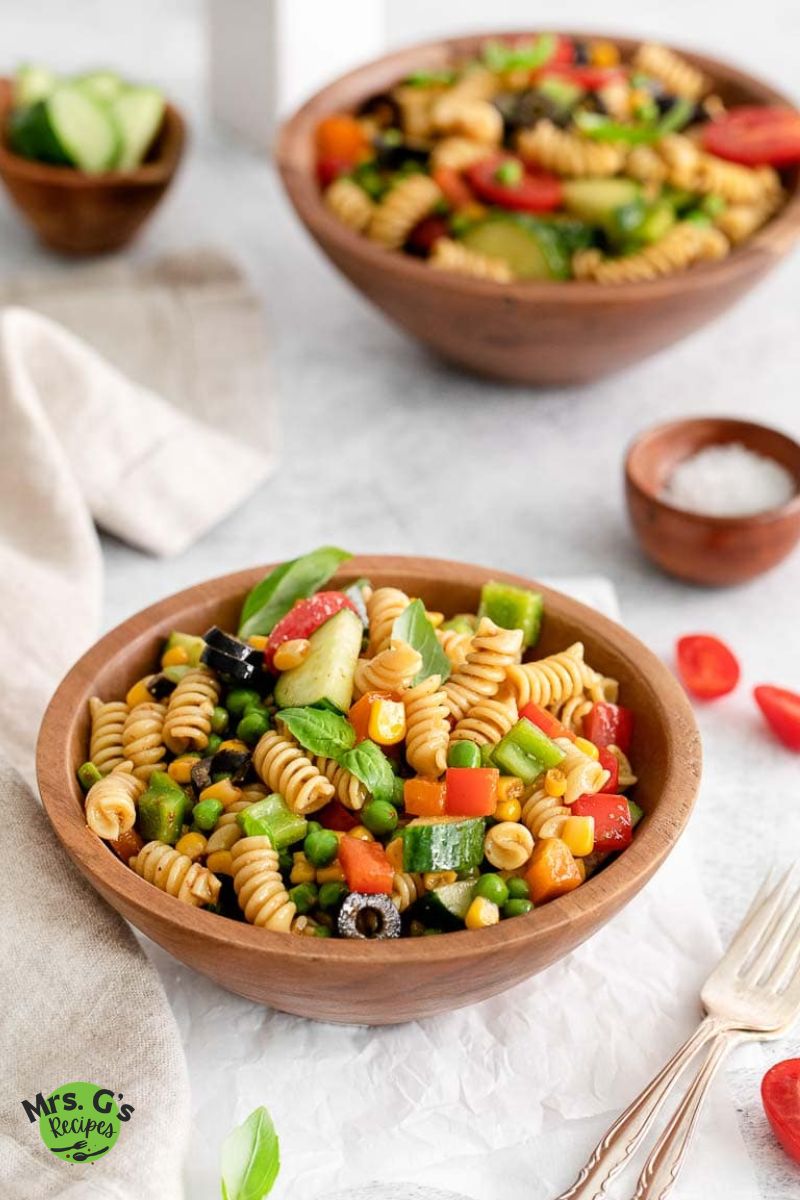 zesty Italian pasta salad and silverwares on a wood bowl with another bowl of salad and salt, cucumber, and cherry tomatoes placed on a white cloth.