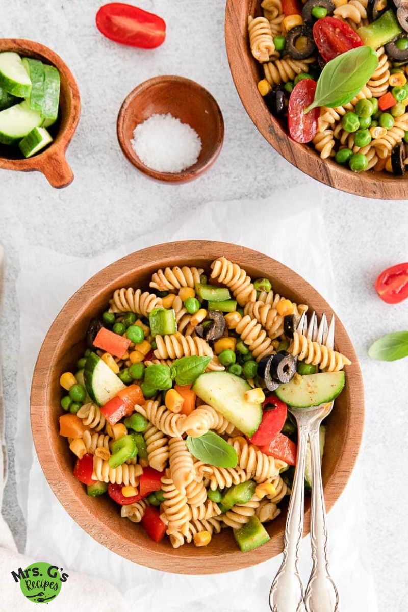rainbow rotini pasta salad and silverware in a wood bowl with another bowl of salad with cucumber, and cherry tomatoes vegetables placed on a white cloth.