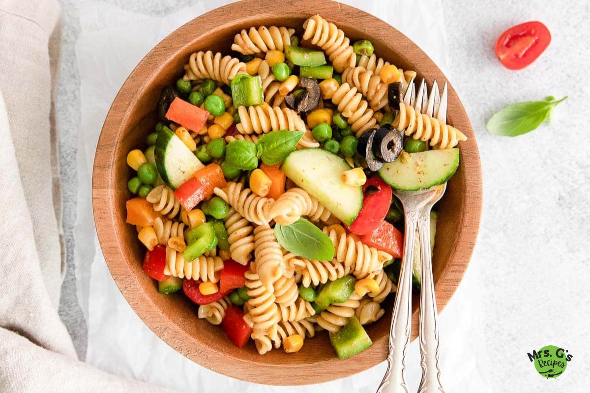 rainbow pasta salad and silverwares on a wood bowl with another bowl of salad and salt, cucumber, and cherry tomatoes placed on a white cloth.